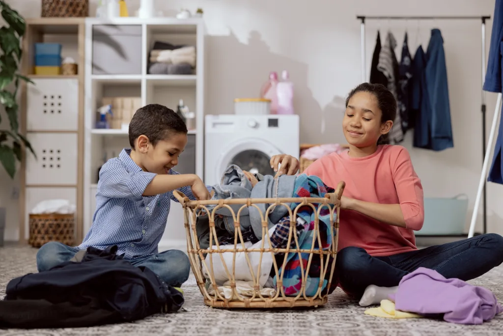 Niños sonrientes ayudando a doblar ropa en casa, en un ambiente ordenado y alegre.